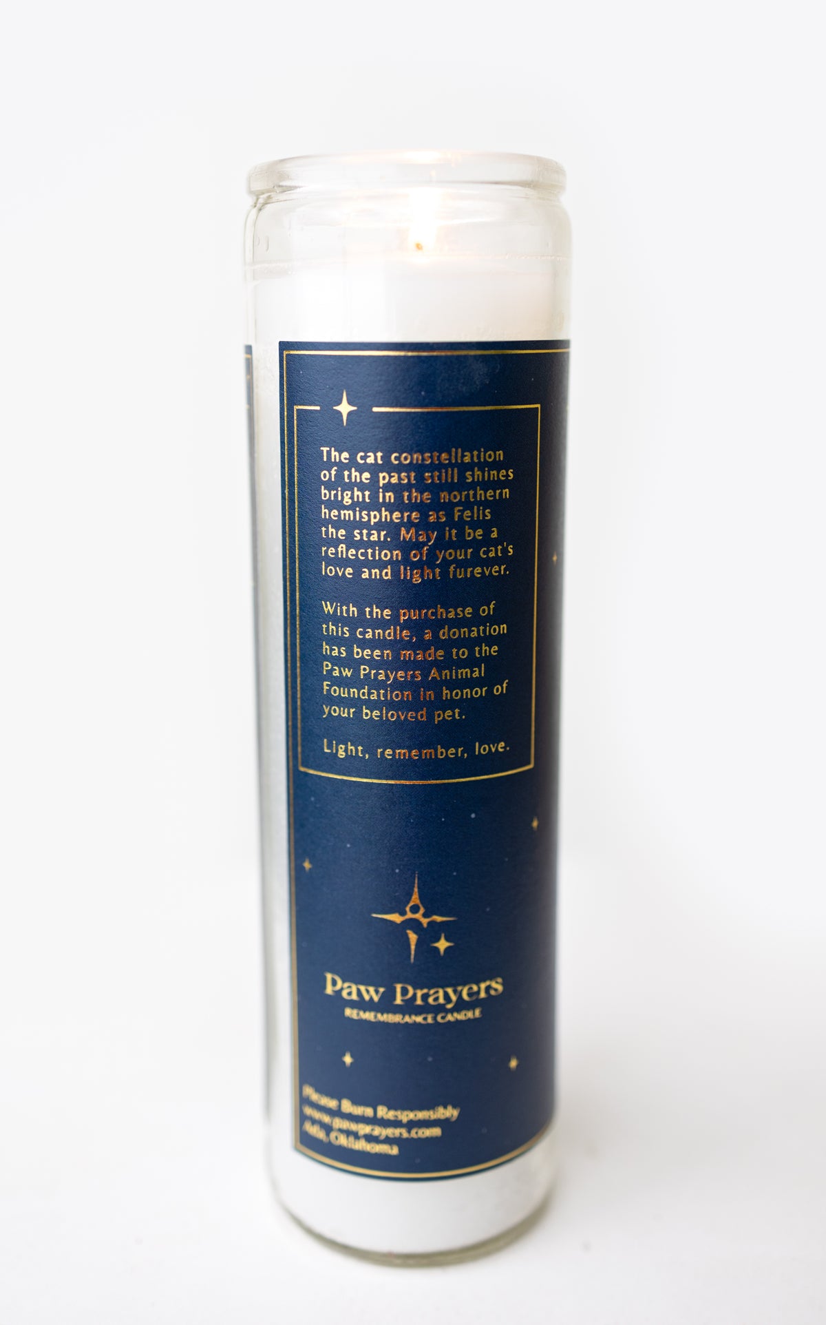 A tall glass prayer candle with white wax and a navy blue label featuring gold text and star illustrations. The label reads 'Paw Prayers' and includes a message about cats and a Paw Prayers Animal Foundation.