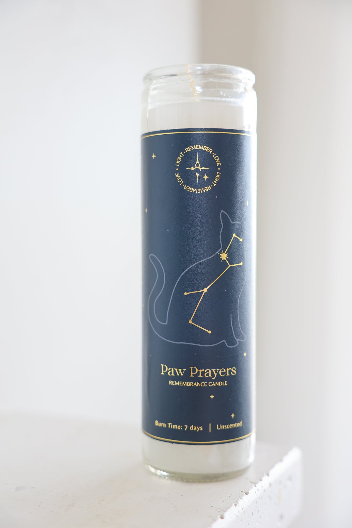 A tall glass jar candle with a navy blue label featuring a white cat constellation design and gold text reading 'Paw Prayers' and Light Remember Love. The candle is photographed upright on a light surface against a soft white background.