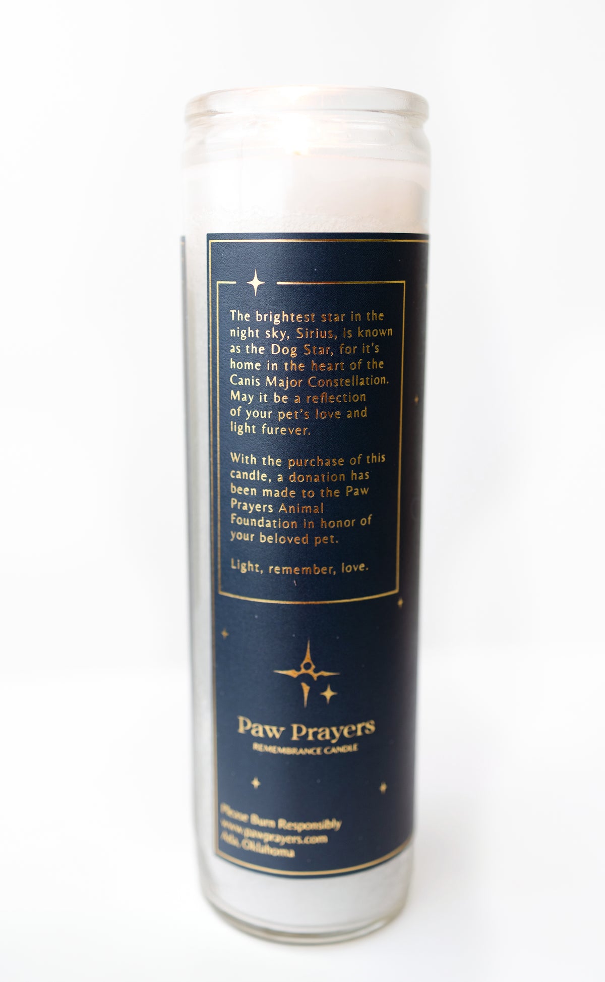 A tall glass jar candle with white wax and a dark navy blue label featuring gold text and star graphics. The label reads 'Paw Prayers Remembrance Candle' and includes a memorial message.