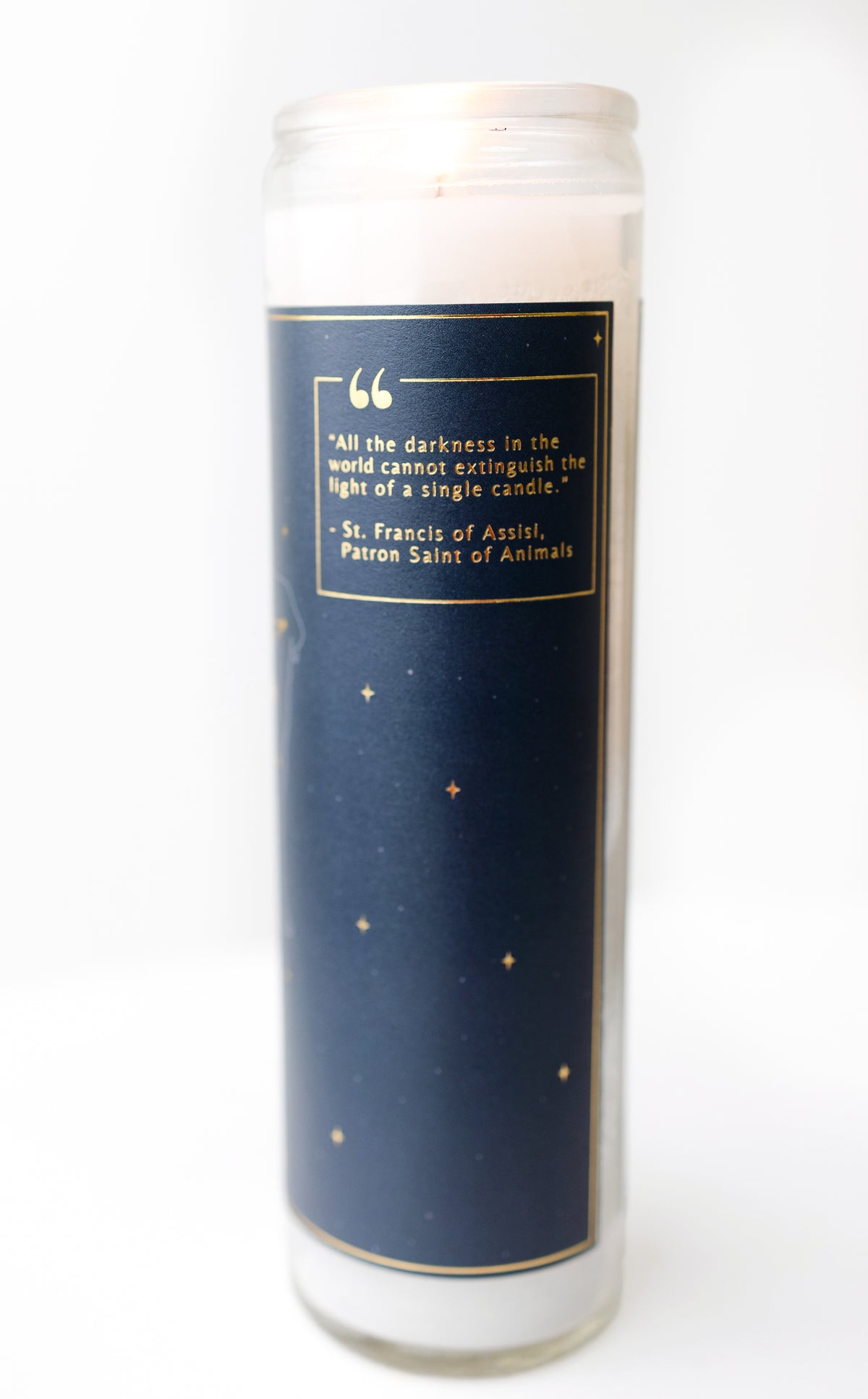A tall glass prayer candle pet memorial git with white wax and a navy blue label featuring gold stars and a quote by St. Francis of Assisi, shown against a plain white background.
